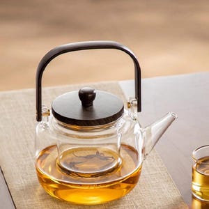May include: A clear glass teapot featuring a dark brown wooden lid with a matching knob and a dark brown arched handle. The teapot contains a removable glass infuser basket and is filled with amber-colored tea, accompanied by a small glass cup.
