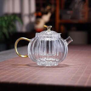 May include: A clear glass teapot with a ribbed, pumpkin-like texture, featuring a gold-colored handle and a small gold-toned knob on the lid. The teapot includes an internal glass infuser basket and a short, straight spout for pouring tea.