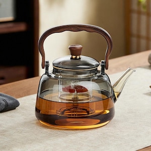 May include: A glass teapot with a wooden handle and lid. The teapot has a built-in infuser and is filled with tea. The tea is a rich amber colour. The teapot is on a light-coloured surface.