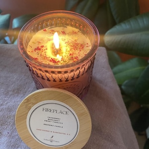 May include: A lit, pink-tinted glass candle with a wooden lid. The candle is labeled "Fireplace" and lists cranberry, bergamot, and sweet tobacco scents. The candle is decorated with dried herbs and flowers.
