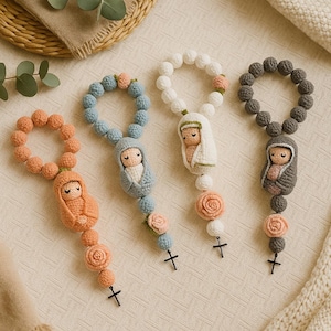 Può includere: Quattro rosari all'uncinetto fatti a mano nei toni del pesca, blu, bianco e grigio. Ogni rosario presenta una piccola figura di bambola, perline decorative e un ciondolo a forma di croce. I rosari sono disposti su una superficie strutturata color crema.