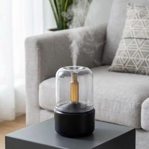 May include: An essential oil diffuser with a black base, a clear glass dome, and a gold-coloured internal component. The diffuser is emitting a stream of vapour. It sits on a dark grey table, with a grey sofa and a plant in the background.