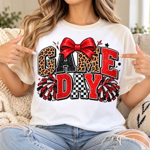 Può includere: Una t-shirt bianca con una stampa grafica che riporta il testo GAME DAY in lettere leopardate, nere e glitterate rosse. Un grande fiocco rosso si trova sopra il testo, affiancato da due pon-pon rossi, bianchi e neri sui lati.
