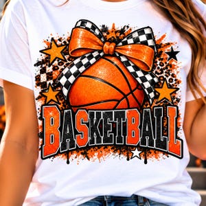 Könnte beinhalten: Ein weißes T-Shirt mit einer Grafik eines orangefarbenen Basketballs, der mit einer schwarz-weiß karierten Schleife verziert ist. Das Design enthält orangefarbene Sterne, Leopardenmuster-Akzente und das Wort BASKETBALL in fetter Schrift.