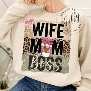 Könnte beinhalten: Cremefarbenes Sweatshirt mit den Worten "WIFE MOM BOSS" in einem mehrschichtigen Design. Das Design umfasst eine Kaffeetasse, rosa Schleifen sowie Blumen- und Leopardenmuster-Akzente. Das Sweatshirt hat eine lockere Passform.