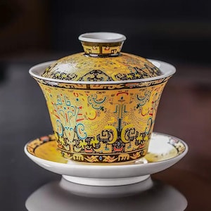 May include: A yellow ceramic tea set with a lid, cup, and saucer. The set features intricate black and colorful patterns on a yellow background. The cup and saucer are white on the inside and have a glossy finish.