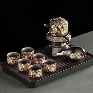 May include: A ceramic tea set with a dark brown tray. The set includes six small cups, a teapot, and a serving pitcher. The cups and teapot are decorated with gold and red butterfly patterns.