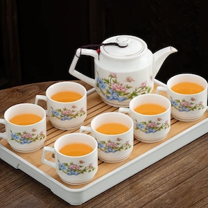 Puede incluir: Juego de té de porcelana blanca con diseños florales, que incluye una tetera y seis tazas llenas de té. El juego está dispuesto sobre una bandeja de bambú. El té es de color naranja dorado.
