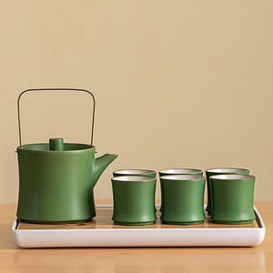 Puede incluir: Un juego de té de cerámica verde con una tetera y seis tazas a juego. La tetera tiene un asa y un pico, y las tazas son cilíndricas. El juego se exhibe en una bandeja blanca con una inserción de estilo bambú, ideal para servir té.