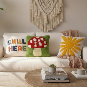May include: A white sofa with three decorative throw pillows. One pillow has the words "CHILL HERE" in colorful letters. Another pillow features a red mushroom design with white spots. The third pillow has a yellow sun design. A macrame wall hanging is visible.