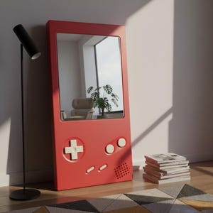 May include: A red mirror designed to resemble a retro handheld games console. It features white buttons and a directional pad. The reflection shows a room with a chair, plant, and window. A black floor lamp and books are also visible.