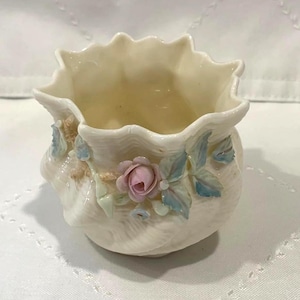 May include: A small, off-white ceramic vase with a ruffled rim. The vase is decorated with a pink rose and light blue leaves. The vase has a textured surface and is likely for decorative purposes.