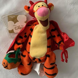 May include: A plush Tigger toy dressed in a red devil costume, complete with a cape and horns. The orange and black striped tiger holds a pumpkin-shaped treat basket and a small trident. A Disney Store tag is attached.