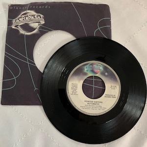 May include: A black vinyl 45 rpm record with a label that says "Pointer Sisters Automatic" and "Produced by Richard Perry". The record is on a white surface with a black and white paper sleeve that says "Planet Records" and has a spaceship logo.