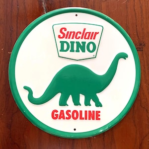 May include: Round metal sign with a white background and a green border. The sign features a green dinosaur silhouette and the words "Sinclair DINO" and "GASOLINE" in red and green text. The sign has two small holes for hanging.