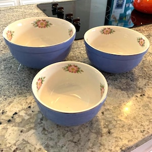 May include: Three ceramic bowls with a light blue exterior and a white interior. Each bowl features a floral design with pink and yellow flowers. The bowls are arranged on a speckled countertop.