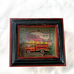 May include: A framed vintage toy fire truck. The red and yellow fire truck is set against a backdrop with a vintage illustration of a fire truck and a boy. The frame is black and red, with the text "FLOOR TOYS" and "MECHANICAL TOYS".
