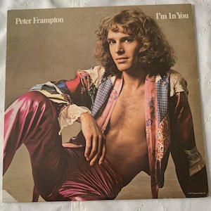 May include: Album cover for Peter Frampton's "I'm In You." The album cover features a man with long, curly hair, wearing a patchwork jacket and shiny, magenta pants. The album title is in the upper right corner.