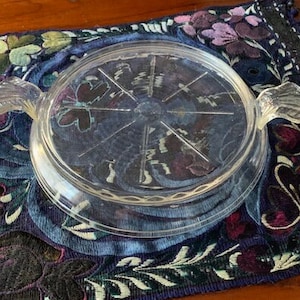 May include: A clear glass hot pad with two handles. The hot pad has a circular design with a raised center and a textured rim.