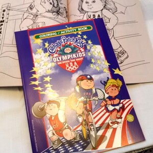 May include: A Cabbage Patch Kids Olympikids 1996 Official Team Mascot USA coloring and activity book with a blue cover featuring illustrations of characters participating in sports, including weightlifting, cycling, and tennis, with yellow stars.
