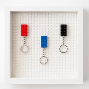 May include: Three rectangular building block keychains in red, blue, and black, each attached to a silver metal split ring. The keychains are mounted on a white square base plate with a stud pattern, enclosed within a white square frame.