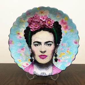 Puede incluir: Plato decorativo con un retrato de Frida Kahlo. El plato tiene un borde festoneado y un fondo azul claro con detalles florales. Kahlo está representada con una corona de flores, una diadema verde y lápiz labial rosa. Pendientes negros completan el look.