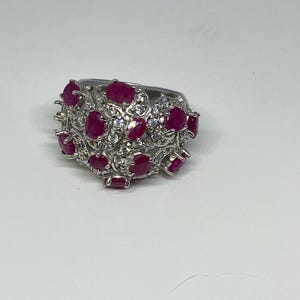 Könnte beinhalten: Ein silberner Ring mit einem Cluster aus ovalen und rechteckigen roten Edelsteinen, durchsetzt mit kleineren, klaren Steinen. Der Ring hat ein verziertes, strukturiertes Design, wobei die Edelsteine in einem blumen- oder rankenartigen Muster gesetzt sind.