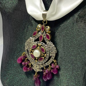 Può includere: Un pendente d'argento ornato con una perla centrale, circondata da pietre rosse e piccole pietre trasparenti. Il pendente è appeso a un nastro di raso bianco. Il design presenta un'intricata lavorazione del metallo e perline pendenti color rubino.