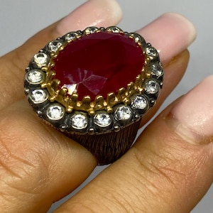 May include: Oval ruby ring with a gold setting and a halo of small, clear gemstones. The ring features a textured, dark metal band. The ruby is a deep red colour, and the gemstones sparkle.