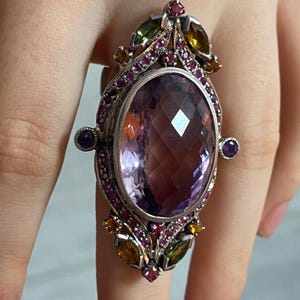 Op de afbeelding: Een sierlijke zilveren ring met een grote, facetgeslepen ovale amethist edelsteen. De ring is versierd met kleinere edelstenen in rood, groen en geel, en heeft een gedetailleerd, bloemachtig ontwerp.