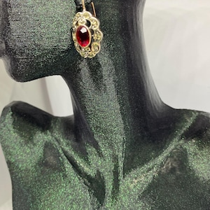 May include: A gold-toned earring with a large, oval, deep red gemstone at its center. The gemstone is surrounded by a halo of smaller, clear stones. The earring is displayed on a black mannequin.