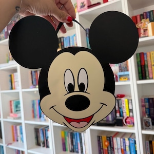 Op de afbeelding: Een zwarte en crèmekleurige Mickey Mouse-vormige tas met zwarte cirkelvormige oren en een zwarte band. De tas heeft een lachend Mickey Mouse-gezicht met een rode mond en zwarte ogen. De tas wordt omhoog gehouden voor een witte boekenkast vol boeken.