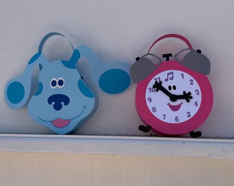 May include: A blue dog-shaped cutout with floppy ears and a smiling face next to a pink alarm clock-shaped cutout with a smiling face, black clock hands, and a musical note symbol on the clock face, both featuring a flat, layered design.