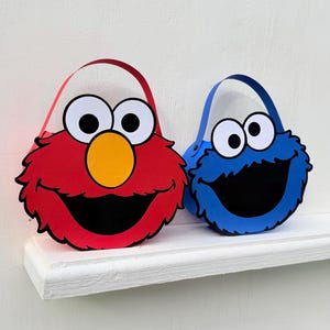 May include: Two colorful, cartoon-themed baskets. One is red with a yellow nose and the other is blue. Both have large eyes and black mouths. Each basket has a matching handle.