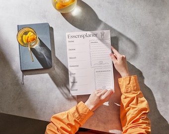 May include: A white meal planner with the German text "Essensplaner" is held by a person wearing an orange shirt. A blue notebook and a glass of drink with a gold straw are on the table.