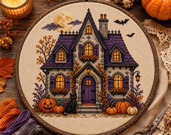 May include: A circular embroidery hoop containing a cross-stitch design of a two-story house with a purple roof, yellow glowing windows, and a purple front door. A black cat sits near the entrance, surrounded by orange pumpkins and autumn foliage.