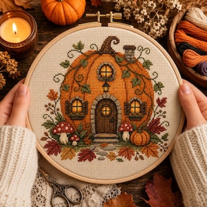 May include: A cross-stitch embroidery project in a circular wooden hoop featuring a large orange pumpkin shaped like a house with a brown door, windows, a chimney, and a lantern. The design includes red and white mushrooms and autumn leaves.