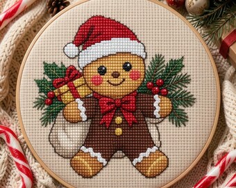 Può includere: Un telaio da ricamo circolare in legno con un motivo a punto croce di un omino di pan di zenzero con cappello da Babbo Natale rosso e papillon rosso. La figura tiene un regalo giallo circondata da aghi di pino verdi e bacche rosse.
