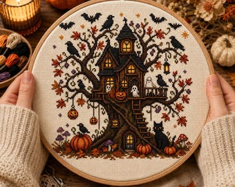 May include: A circular wooden embroidery hoop containing a cross-stitch pattern of a dark treehouse with glowing windows, surrounded by autumn leaves, black crows, bats, ghosts, pumpkins, and a black cat on a light beige fabric background.