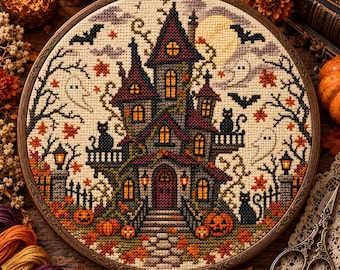May include: A circular embroidery hoop containing a cross-stitch pattern of a multi-storey haunted house. The design features a dark mansion with glowing windows, black cats, ghosts, bats, pumpkins, and autumn leaves against a pale background.