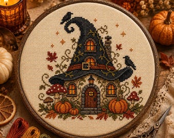 Può includere: Un telaio da ricamo circolare contenente un disegno a punto croce di una casa a forma di cappello da strega. Il design presenta un tetto a cappello blu scuro, una porta marrone, finestre arancioni, zucche, funghi rossi e uccelli neri su tessuto color crema.