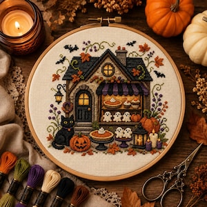 May include: A circular wooden embroidery hoop containing a cross-stitch pattern of a house with a black cat, pumpkins, ghosts, and autumn leaves. The design features a storefront with pies and treats, surrounded by small bats and purple floral accents.