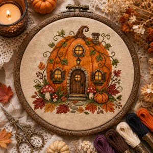 May include: A cross-stitch embroidery in a circular wooden hoop featuring a large orange pumpkin shaped like a house with a door, windows, and a chimney. The design includes green vines, red and orange autumn leaves, and small red and white mushrooms.