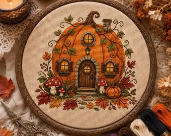 May include: A cross-stitch embroidery in a circular wooden hoop featuring a large orange pumpkin shaped like a house with a door, windows, and a chimney. The design includes green vines, red and orange autumn leaves, and small red and white mushrooms.