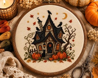 Può includere: Un telaio da ricamo circolare in legno contenente un motivo a punto croce di una casa scura con finestre arancioni, corvi neri, una luna crescente e zucche. Il disegno presenta foglie autunnali, un gatto nero e un albero spoglio su tessuto crema.