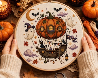 May include: A circular wooden embroidery hoop containing a cross-stitch pattern of a pumpkin-shaped hot air balloon. The design features a ghost, a black cat, a raven, a crescent moon, bats, purple clouds, and autumn leaves on a cream fabric.