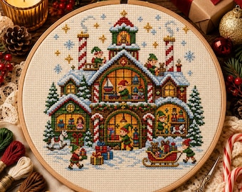 May include: A circular wooden embroidery hoop containing a cross-stitch pattern of a multi-story workshop covered in snow. The scene features elves, toys, gift boxes, candy cane pillars, and evergreen trees, all rendered in vibrant, festive colors.