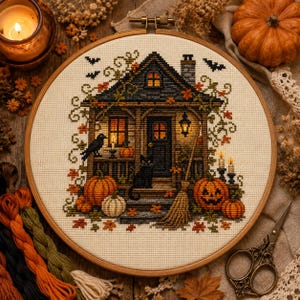 May include: A circular embroidery hoop containing a cross-stitch pattern of a dark house with a black cat, a raven, pumpkins, and a broom. The scene features autumn leaves, lit windows, and flying bats on a cream-colored fabric background.
