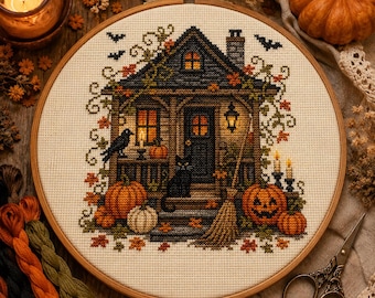 May include: A circular embroidery hoop containing a cross-stitch pattern of a dark house with a black cat, a raven, pumpkins, and a broom. The scene features autumn leaves, lit windows, and flying bats on a cream-colored fabric background.