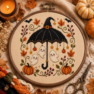May include: A circular wooden embroidery hoop containing a cross-stitch pattern on beige fabric. The design features a black witch hat umbrella with three white ghosts, orange pumpkins, autumn leaves, and small stars in black, orange, and green.